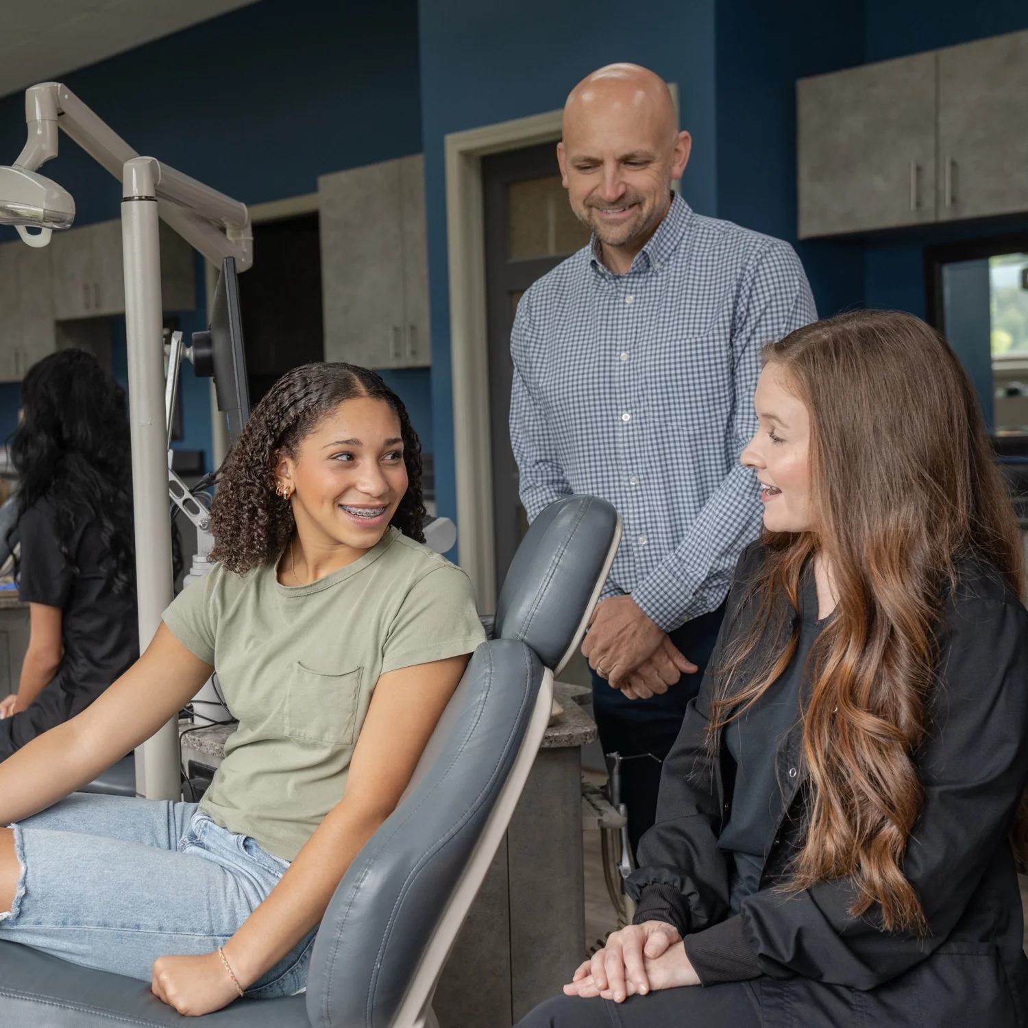 A teenage girl with curly brown hair sits in a gray dental chair. She is smiling, looking off to the side, and has metal braces on her teeth. She is wearing a light green t-shirt with a pocket and blue jeans. In the background, a person in a plaid shirt is partially visible, along with a blue wall and medical equipment.