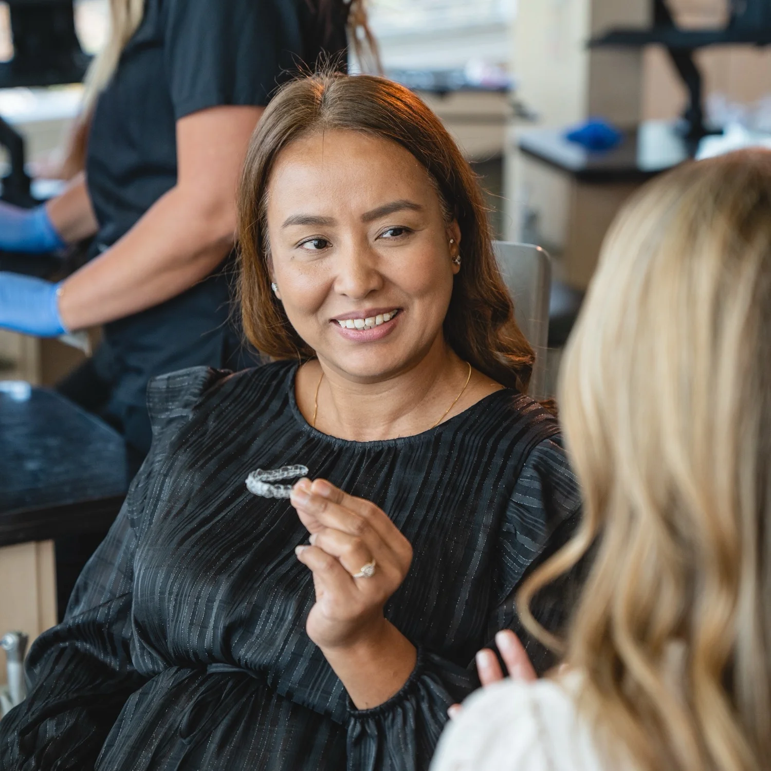 A middle-aged woman with brown hair smiles while holding a clear Invisalign aligner. She is wearing a black, pleated, long-sleeved blouse and a thin gold necklace. She is talking to a person with blonde hair whose back is to the camera. In the background, a person in a black scrub top is partially visible.