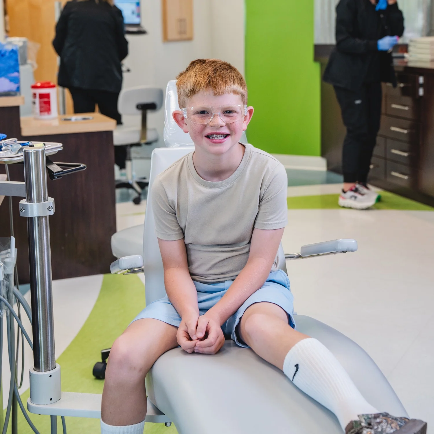 A smiling young boy with reddish-blonde hair and freckles sits in a dental chair. He is wearing clear safety glasses and has metal braces on his teeth. He is wearing a light tan t-shirt and blue shorts. The background is a brightly lit orthodontic office with a green wall.